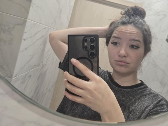 Emeline Minacucci, com o cabelo preso em um coque bagunçado, segurando um telefone preto com uma capa de proteção, vestindo uma camiseta cinza, olhando para o espelho, levantando a mão atrás da cabeça; o reflexo no espelho mostra seu rosto e o telefone..
