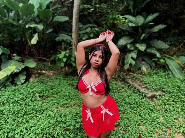 CelesteMorel with brown hair, red bra, and red skirt with white bows, arms raised, standing on green grass, looking at camera, medium shot, forest background.