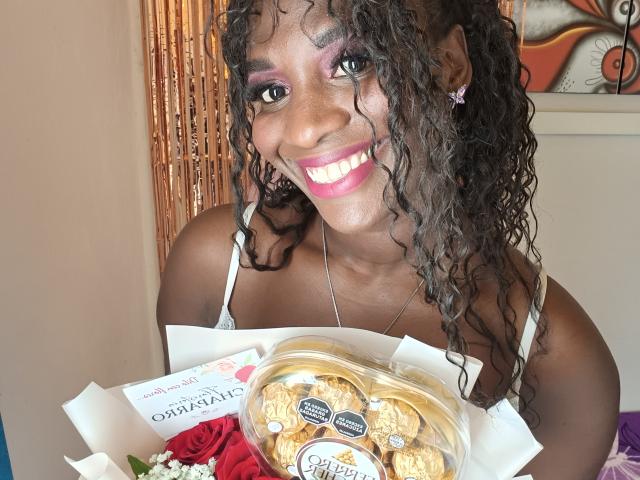 SaraBii, con i capelli ricci scuri, l'ombretto rosa e il rossetto rosa acceso, sorride, tenendo davanti a sé un contenitore di biscotti, indossa un reggiseno bianco e tiene in mano delle rose rosse..