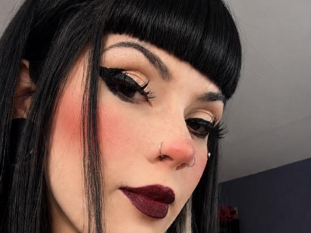 AmyDan69 has black hair with bangs, dark eyeliner, long lashes, pink blush, red lipstick, nose ring, and a septum ring, looking down with a slightly bored expression.