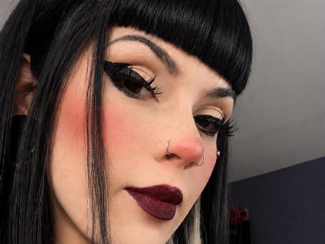 AmyDan69 has black hair, pale skin, bold eyeliner, long eyelashes, red blush, dark lipstick, nose ring, and a nose piercing. She looks down with a slight smile.