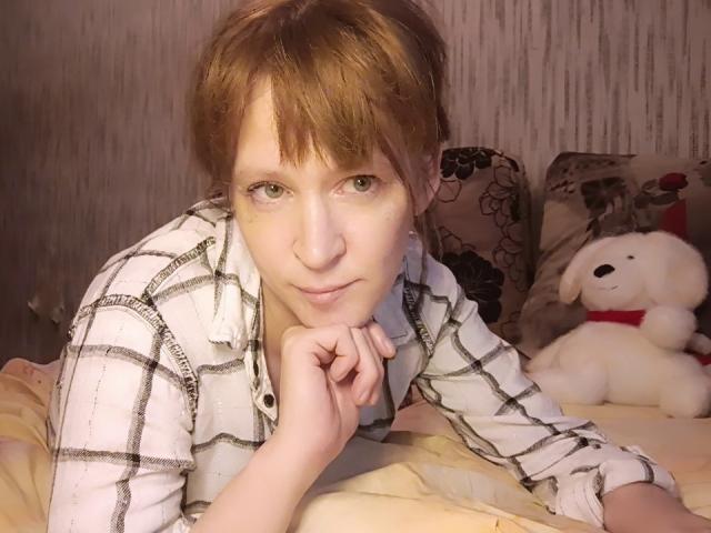 NettyBrittany with red hair, green eyes, and pale skin, wearing a plaid shirt, resting chin on hand, looking at camera, lying on bed with white stuffed dog.
