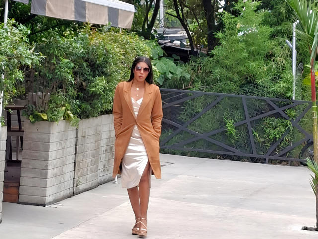 CamilaWest, con un cappotto color caramello, un vestito bianco e sandali con cinturini, in piedi su un vialetto di cemento, indossa occhiali da sole, ha i capelli castani e un'espressione sicura, circondata dal verde..