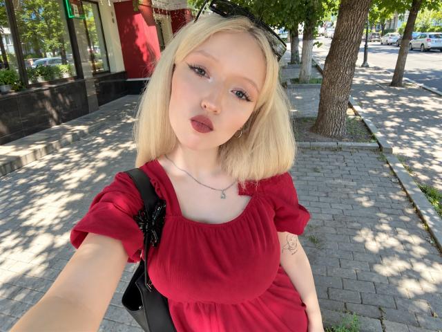 Yael Overman, rubia, con labios carnosos, vestido rojo, pechos pequeños y un tatuaje en el brazo, se está tomando un selfie en una acera soleada, usando gafas de sol negras y un bolso negro..