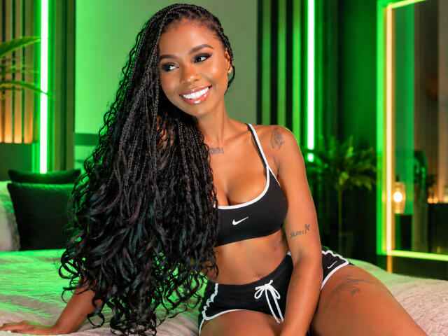 Kinkyandsavage with long black braids, smiling, wearing a black Nike sports bra and black shorts with white trim, sitting on a bed, showing her tits and a tattoo.