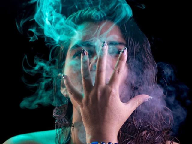 BulmaHot's face is covered in blue-green smoke, her hand with black nail polish in front, dark wavy hair, and a black background. She looks intense and smoky.