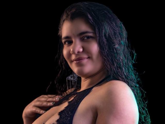 BulmaHot with wet black lace top, smiling, showing her big boobs, dark wavy hair, hand on chest, against black background. Soft lighting, greenish-blue highlight.