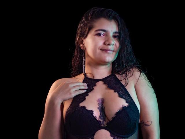 BulmaHot with wet dark hair, smiling, wearing a black lace bra that reveals her tits and a tattoo on her right arm, against a black background.