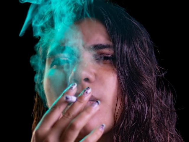 BulmaHot with dark, wavy hair smoking a cigarette, blue-green smoke swirling around her face, hand with painted nails holding the cigarette, black background.