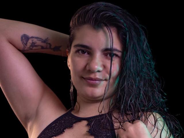 BulmaHot with wet black hair, arms raised, showing a fish tattoo on her right arm, wearing a black lace top, smiling slightly. Her tits are partially visible.