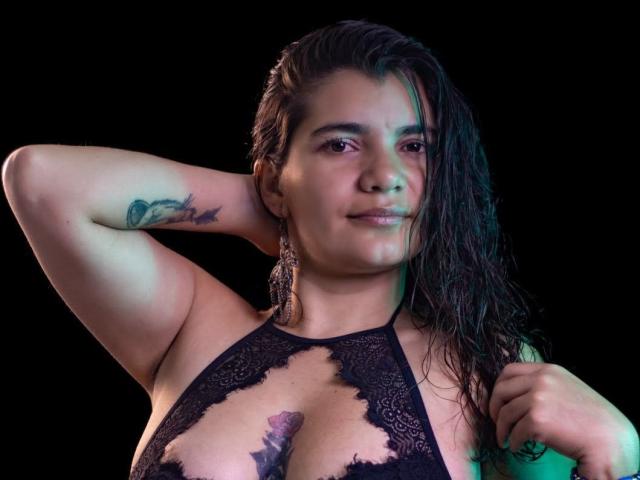 BulmaHot with wet dark hair, wearing a black lace top, showing her big tits and a tattoo on her arm, looking at the camera with a slight smile.