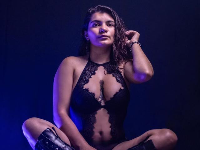 BulmaHot with wet brown hair, wearing a black lace bodysuit, sitting cross-legged, showing her tits, in knee-high black boots, against a blue-lit background.