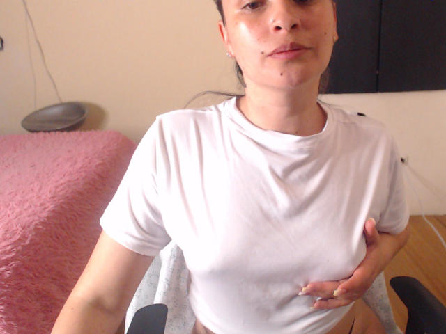 WendySchulz with a slight smile, wearing a white shirt, hand on her chest, showing her small tits, pink bed in the background, black chair, and wooden floor.