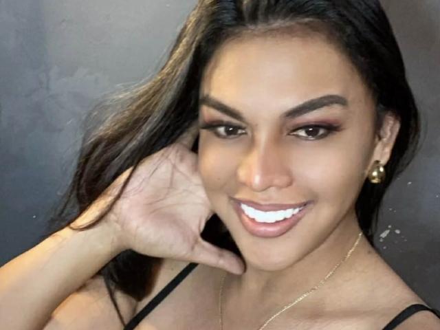 Un selfie de CummingSoon con cabello oscuro, ojos marrones y una gran sonrisa blanca. Está tocándose la cara, lleva aretes dorados y un top negro con tirantes..