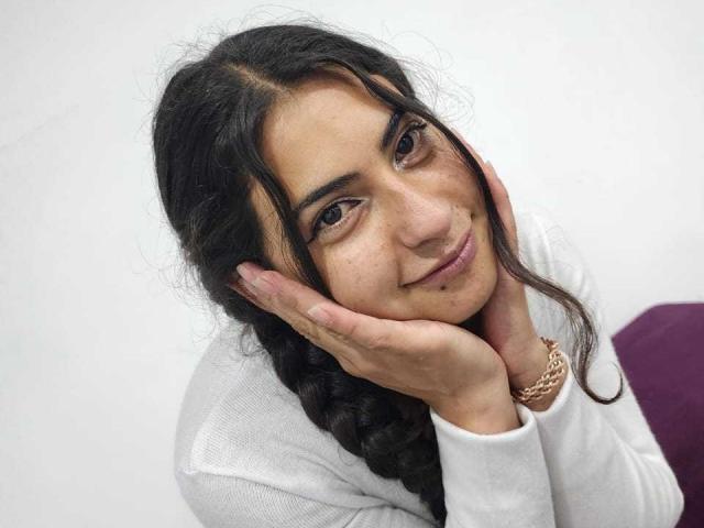 AngelAlba with dark, wavy hair in a braid, hands on cheeks, soft smile, brown eyes, wearing a white sweater, gold bracelet, and a purple pillow.