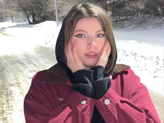 LoriKai with hands on cheeks, wearing a red jacket with brown collar, black hood, and black sleeves, standing in a snowy landscape, brown eyes, light skin, and brown hair.