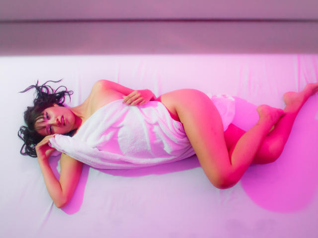 HanaJoud lying on white bed, naked, covered by white towel, pink and purple light, black curly hair, pale skin, small tits, bare ass, relaxed, hand under head, legs crossed.