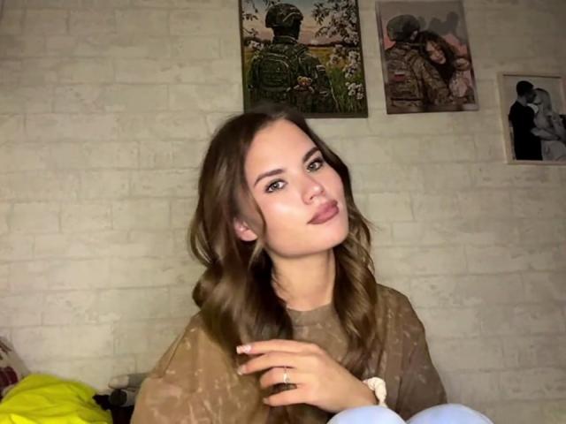 SheritaMerola with wavy brown hair, pouty lips, and ring on right hand, wearing a brown top, hands together, in a dimly lit room with paintings on a white brick wall.