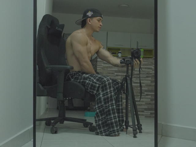 NoahhJacob, shirtless, muscular, wearing black cap and plaid pajama pants, sitting on chair, holding camera on tripod, white room, mirror reflection.