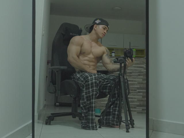 NoahhJacob, shirtless with a muscular build, wearing plaid pajama pants and a backward baseball cap, sitting in a chair holding a camera, reflected in a mirror.