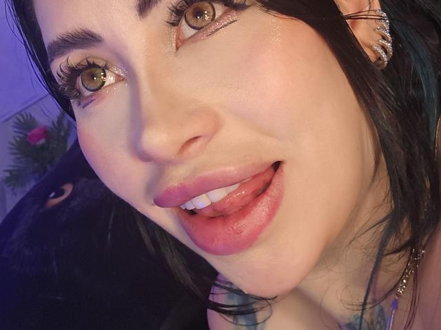 Kkole's face with green eyes, pink glossy lips, tongue slightly out, black hair, and a nose piercing. She's wearing earrings and has a tattoo visible on her neck.