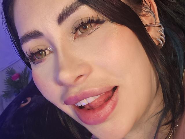 Kkole's face, brown eyes, long eyelashes, glossy pink lips, tongue slightly out, dark hair, silver earrings, glowing skin, subtle makeup, smiling, close-up, looking at camera.
