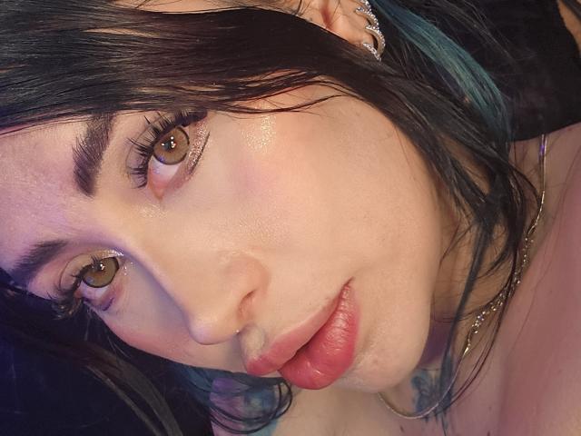 Kkole's face, showing brown eyes, glossy pink lips, and black hair with teal highlights, wearing a nose ring and ear gauges, with a slight smile and shiny skin.