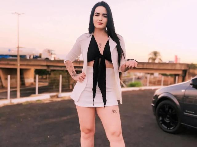 YaneBranquinha mit langen schwarzen Haaren, einer weißen Jacke, einem schwarzen, gerafften Oberteil und einem weißen Minirock, steht selbstbewusst mit den Händen in den Hüften, ein schwarzes Auto ist rechts zu sehen, ein Tattoo auf dem rechten Oberschenkel..