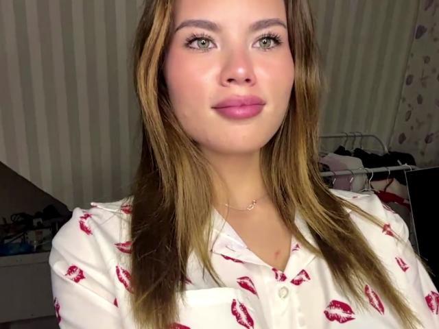 SheritaMerola with light brown hair, green eyes, pink lipstick, wearing a white shirt with red kiss prints, necklace, standing in a room, looking at camera, slight smile.