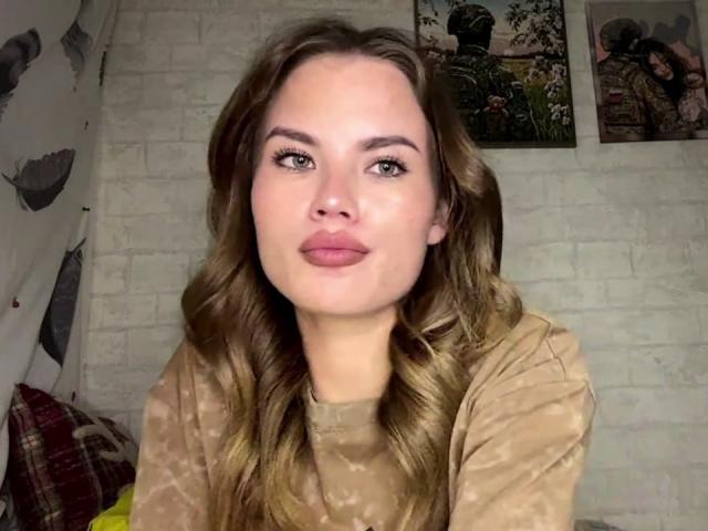 SheritaMerola with wavy brown hair, light skin, and full pink lips, wearing a beige tie-dye shirt, looking into the camera with a slight smile.