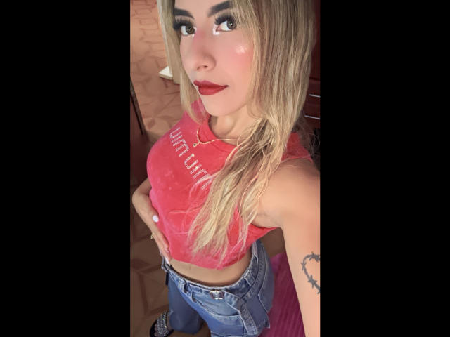 CloeRouss with blonde hair, red lipstick, and a pink crop top showing her tits, wearing blue jeans, standing with hands on hips, looking up at the camera.