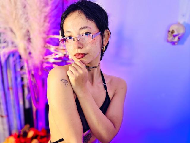 Roxannee with short black hair, glasses, red lipstick, freckles, black halter top, choker, tattoo on left shoulder, holding collar, purple-blue lighting, puffy pink plant on left.