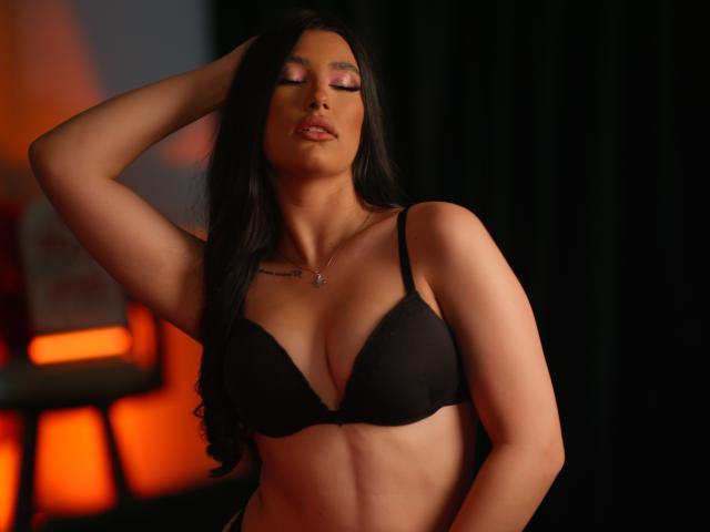 NeraRose with dark hair, eyes closed, wearing a black bra, hand in hair, soft orange light, tan skin, necklace, small tattoo above cleavage, sexy, confident, slightly parted lips.