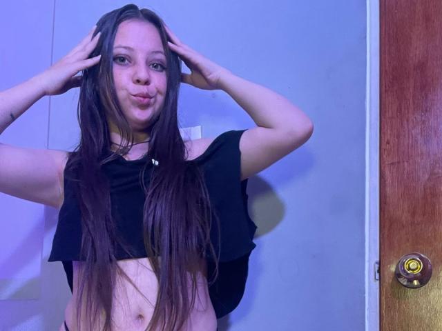GraceAndTony with long brown hair, pouty face, hands in hair, black crop top, showing her small tits and belly button, standing in front of a wooden door.