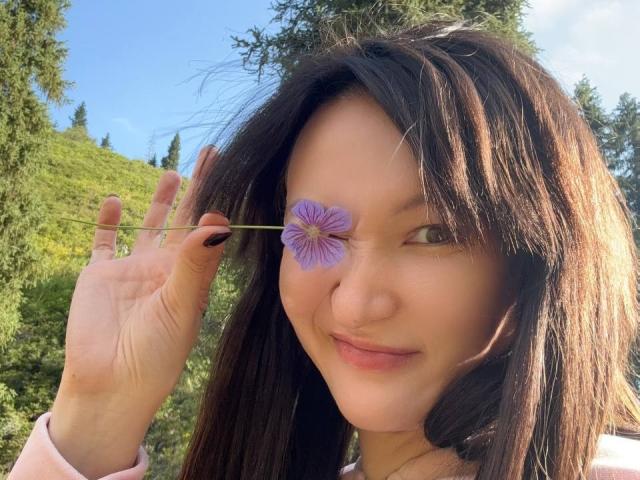 LanaJensenn sosteniendo una flor morada sobre su ojo derecho, sonriendo, con cabello castaño oscuro, piel clara y esmalte de uñas negro, en un entorno exterior soleado..