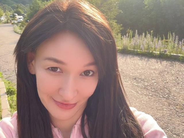 LanaJensenn tem cabelo castanho liso, pele clara e um pequeno sorriso. Ela está vestindo um moletom rosa, e seus olhos castanhos estão olhando para a câmera. A luz do sol ilumina seu rosto..
