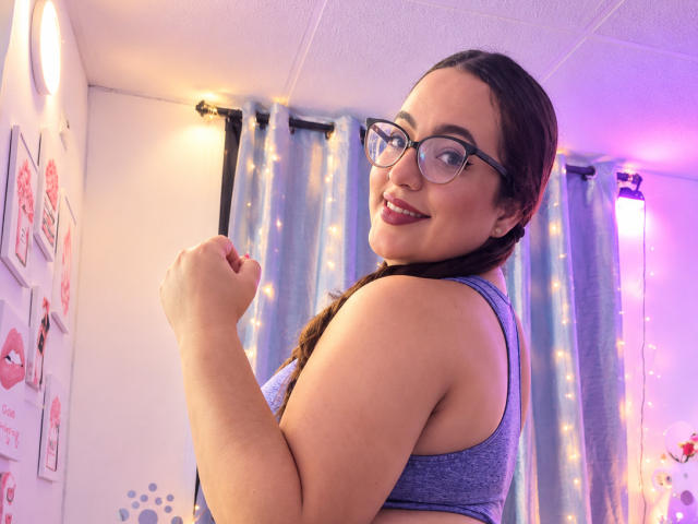 StacyMillerr with glasses, smiling, showing her tits in a blue tank top, braided hair, holding fist up, pink lipstick, white ceiling, blue curtains, and string lights in background.