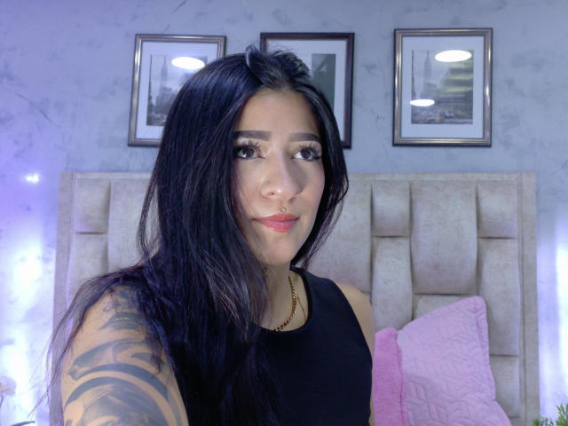 LuciadelaVega with long black hair, black sleeveless top, tattoo on right arm, nose ring, pink pillow, and three framed pictures on a grey wall, smiling at the camera.