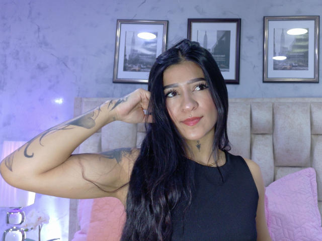 LuciadelaVega with long black hair, black sleeveless top, tattooed arm, nose ring, and pink lipstick, smiling, hand in hair, pink pillow, white headboard, framed pictures in background.