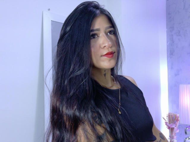 LuciadelaVega with long black hair, red lipstick, nose ring, black sleeveless top, necklace, and tattoo on her neck, looking at the camera with a slight smile.