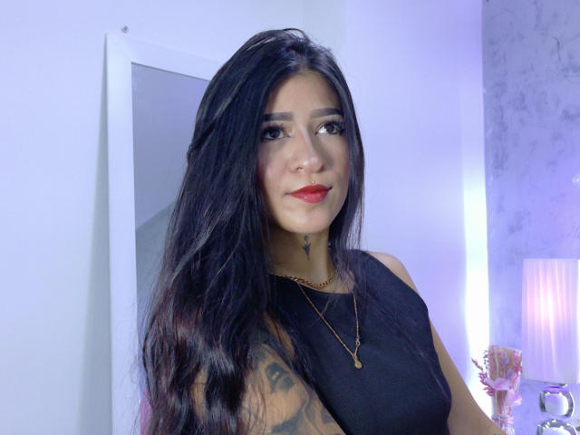 LuciadelaVega with long black hair, red lipstick, black sleeveless top, tattoo on left arm, gold necklace, smiling at camera, white wall, mirror, and purple-lit lamp in background.