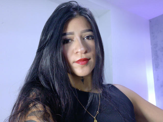LuciadelaVega with long black hair, nose ring, red lipstick, black sleeveless top, and gold necklace, smiling softly, her tits partially visible, in a subtle purple-lit room.