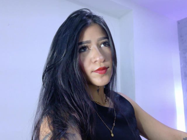 LuciadelaVega with long black hair, brown eyes, nose ring, red lipstick, black sleeveless top, gold necklace, slight smile, and a tattoo on her right arm.