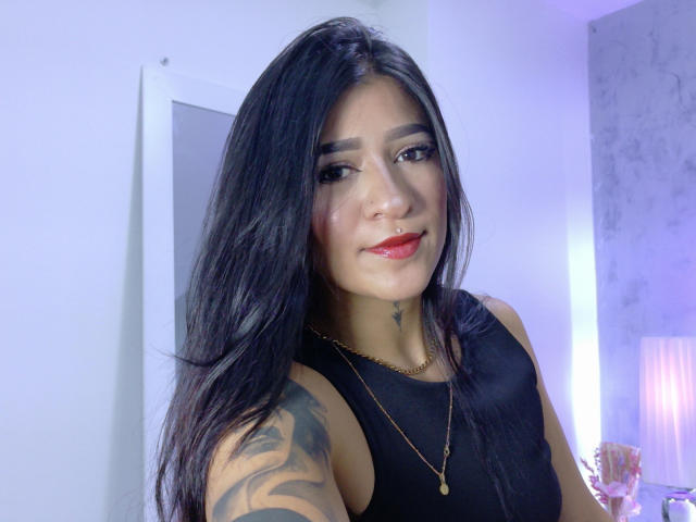 LuciadelaVega with long black hair, red lipstick, black sleeveless top, gold necklace, tattoo on right arm, subtle smile, and a small star tattoo below her chin.