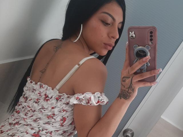RoxannaMoretti with long black hair, white floral dress, off-shoulder, holding pink phone, tattoo on wrist and back, large hoop earrings, looking in mirror, taking selfie.
