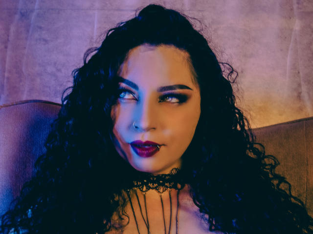 ScarlattaaDesire mit lockigem, schwarzem Haar, dunkelrotem Lippenstift und einer schwarzen Spitzenkette, die zur Seite blickt, mit blau-violetter Beleuchtung auf ihrem Gesicht und ihrer Brust..