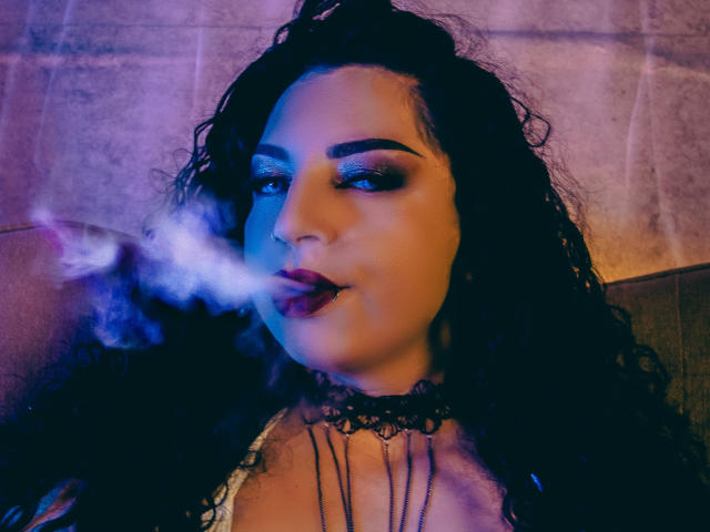 ScarlattaaDesire with curly black hair, smoky blue eyes, and dark lipstick, smoking a joint, exhaling blue-white smoke. Wearing a black lace choker, neon purple and blue lighting.