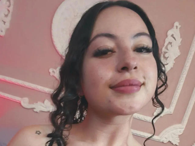 MiiaBennet has wavy black hair, dark eyeliner, and pink lipstick. Her small tits are visible, and she's smiling with a slight pout. A small star tattoo is on her shoulder.