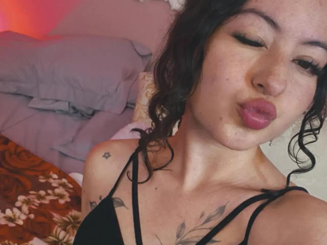 MiiaBennet with pouty pink lips, winking, curly black hair, tattoos on chest and shoulder, wearing black strappy top, slight freckles, cute and sexy.