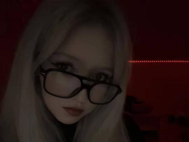 YaelOverman, con la pelle chiara, i capelli lunghi e bianchi, gli occhiali neri e il rossetto rosso, guarda leggermente verso il basso con un sorriso dolce, in una luce rossa tenue..